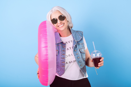 Summer is coming. Glad senior beautiful woman smiling and holding cocktail and ring lifebuoy while standing against isolated blue background.の写真素材