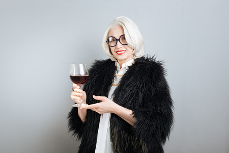Pleasant time. Senior elegant pretty woman smiling and holding wineglass while standing against isolated gray background.の写真素材