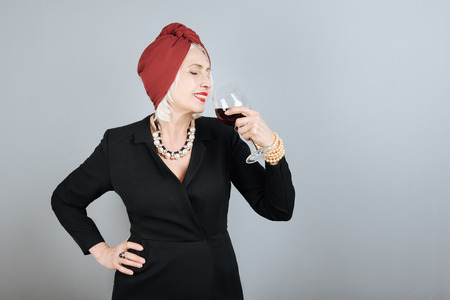 : Good taste. Emotional pretty senior woman drinking wine and smiling while standing against...