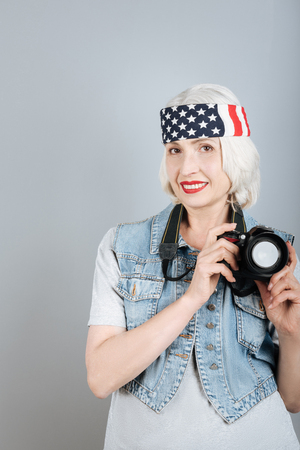 Want to take a picture. Charming senior positive woman holding camera and smiling while standing against isolated gray background.の写真素材
