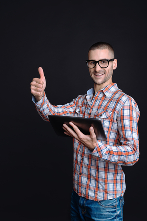 Great job. Young smiling man wearing glasses holding his right thumb up while using tablet computer standing against black backgroundの写真素材
