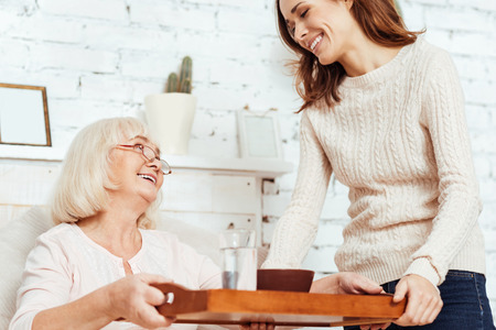 In elated mood. Low angle of pleasant joyful woman holding tray and takign care of her sick grandmother who is lying in bed at homeの写真素材