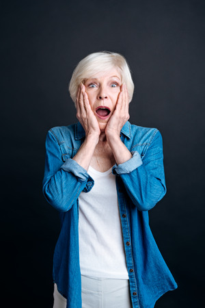 Wow. Astonished elderly woman holding her hands on the fce and lookign at you while being surprisedの写真素材