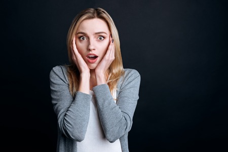 On the edge of emotions. Pleasant emotional young woman holding her hands on the face and looking at you while expressing wonderの写真素材