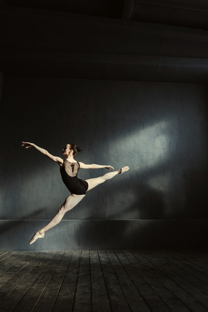 Demonstrating the twine in the air. Flexible masterful principal ballet dancer performing isolated in the black colored room and jumping in the twine while expressing eleganceの写真素材