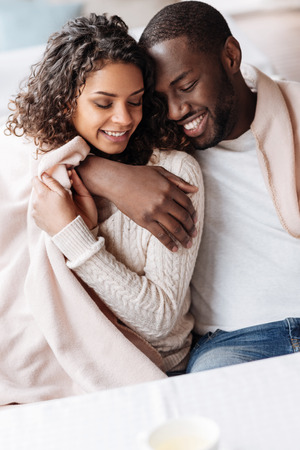 Happy to be in love. Positive young delighted African American couple sitting in the cafe and being covered with a blanket while expressing love and huggingの写真素材