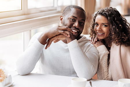 Positive atmosphere around us . Charming positive smiling African American couple having the date in the cafe and being covered with a blanket while expressing happiness and loveの写真素材