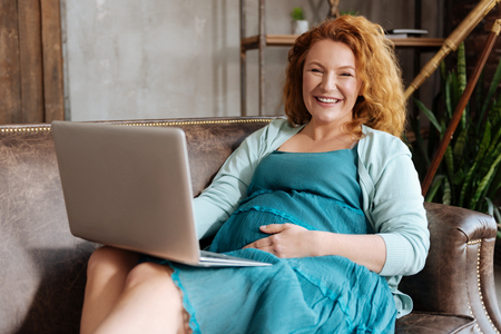 Radiant expectant mother working on laptop on sofaの写真素材