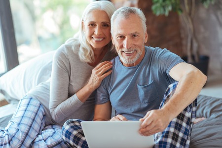 Cheerful aged couple using laptop in bed