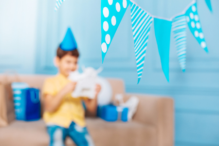Birthday party. The focus being on party flags hanging in the room decorated for birthday party while little boy in party hat sitting on the sofa and examining toy robotの写真素材