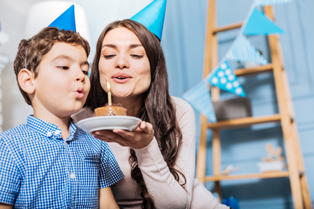 Happy to celebrate. Upbeat young mother holding a plate with a birthday cake and blowing out a candle on it together with her son, being happy to celebrate his birthdayの写真素材