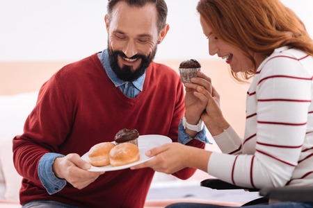 Having a dessert. Joyful bearded man and blond woman laughing and holding together a plate with some cookies and the woman holding the mans handの写真素材