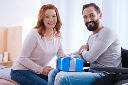 Life is great. Loving and caring woman and a happy disabled man smiling and sitting in a wheelchair while she holding his hand and the man holding a presentの写真素材