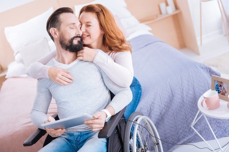 Smiling blond woman of middle age and a bearded paralyzed man holding a tablet while she hugging and caressing him from behind and their eyes closedの写真素材