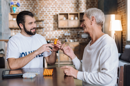 Kind cheerful young social worker recommending useful pills to a smiling senior man while visiting...