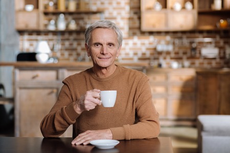 Pleasant man. Pleasant calm cheerful pensioner sitting at the table and looking peaceful while drinking his favorite teaの写真素材