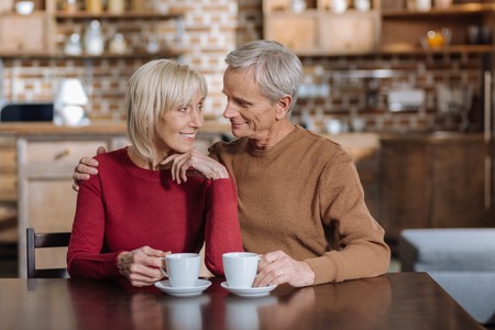 Loving couple. Cheerful smiling aged woman smiling while looking at her romantic aged husband sitting by her side and hugging herの写真素材