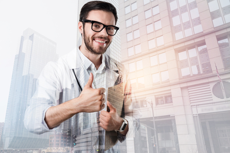 Satisfied doctor. Cheerful positive young doctor smiling and showing his good attitude to work while holding important documentsの写真素材