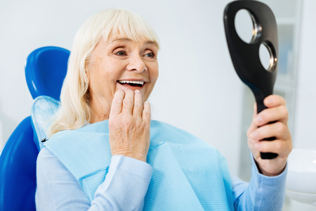 Cannot stop admiring. Close up of cheerful woman looking in the mirror while sitting on the dental chair and expressing positive emotionsの写真素材