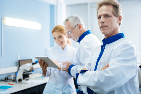 Serious medical researcher of middle age standing in the lab and smiling experienced scientists looking at the screen and wearing uniforms in the backgroundの写真素材