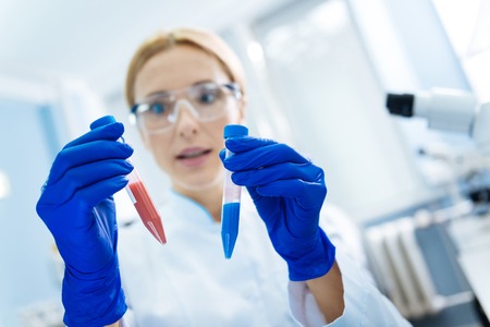 Concentrated promising blond scientist holding test tubes and wearing a uniform and medical gloves...
