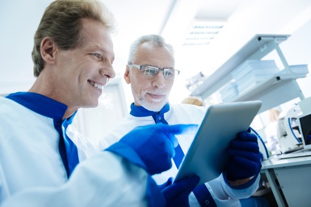 Smiling content grey-haired researcher discussing analysis results displayed on a tablet with a handsome inspired scientist and they wearing uniforms while being in the labの写真素材