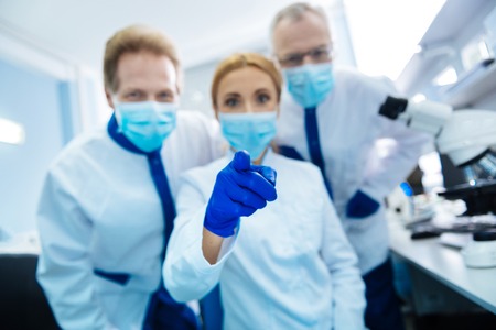 Cheerful vigorous experienced researchers wearing a uniform and a medical face mask and gloves and...