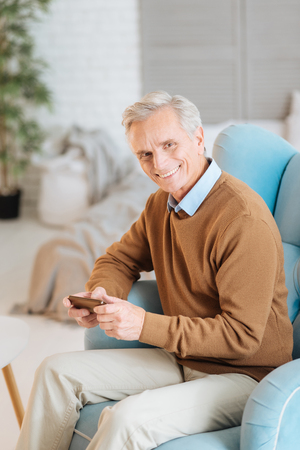Radiant elderly gentleman looking into the camera with a cheerful smile on his face while sitting in a comfy chair and using his phone at home.の写真素材