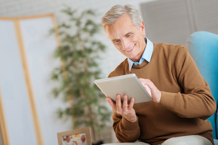 Low angle shot of a retired man sitting in a chair and grinning broadly while using a tablet computer at home.の写真素材