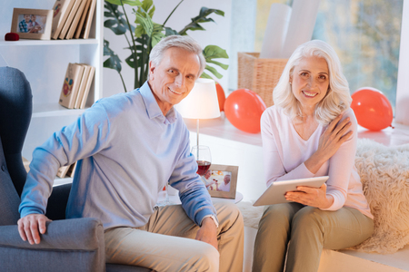 Positive minded senior couple looking into the camera with cheerful smiles on their faces while relaxing at home with a glass of wine and watching videos on a tablet.の写真素材