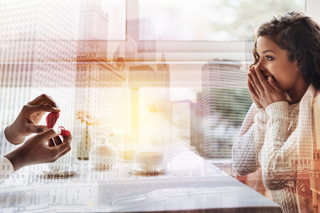 Excited young girl touching her face and expressing amusement while her boyfriend holding a ring and proposing to herの写真素材
