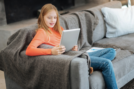 Delighted positive cute girl holding a tablet and using it while resting at homeの写真素材