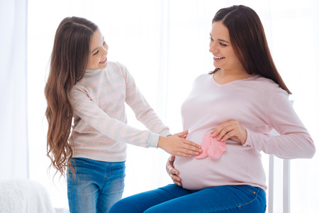 Pleasant happy beaming girl caressing pregnant moms tummy while she holding pink socks and sitting on the chairの写真素材