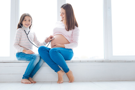 Miracle with stethoscope.  Pregnant woman showing her tummy to surprised fascinated girl  wearing stethoscope  while relaxing on the window sillの写真素材