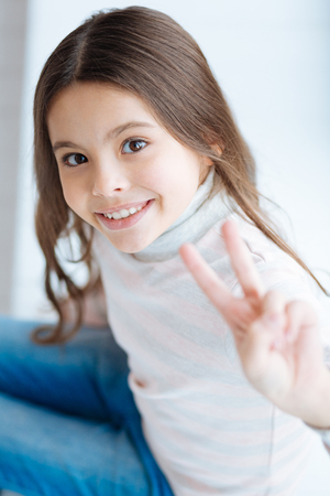 Charming optimistic small girl showing sign of victory while sitting and gazing at the cameraの写真素材