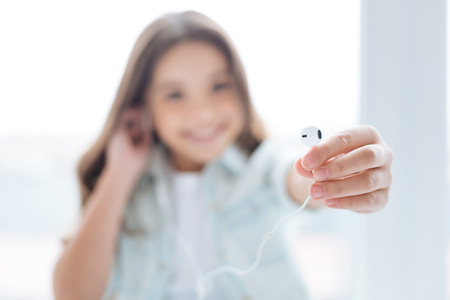 Selective focus of white earphone when brunette little girl stretching it to the camera while...