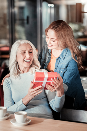 Unexpected present. Happy cheerful emotional senior woman sitting at the table and receiving a red box with a present from her loving attentive relativeの写真素材