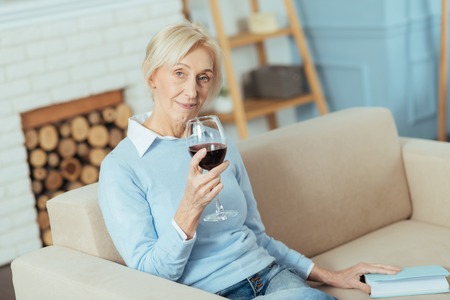 Drinking wine. Peaceful kind aged woman looking relaxed while sitting at home on a soft sofa and...