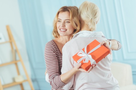 Greeting granny. Smiling kind calm woman looking happy while holding a big beautiful bright present and hugging her cute aged grandmotherの写真素材