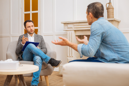 Problem solving. Handsome prosperous male psychologist asking patient about problems while he talking and sitting on the sofaの写真素材