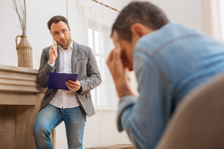 Solving trouble. Handsome thoughtful male psychologist holding blue folder and looking at patient while he cryingの写真素材