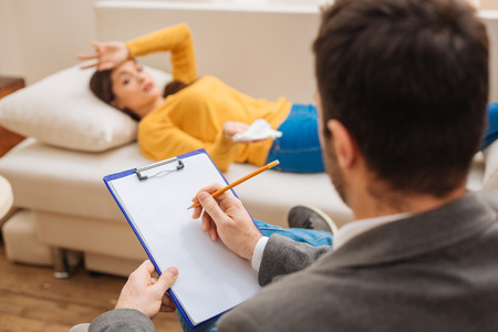 Therapy process. Attentive male psychologist holding folder and pencil while female patient lying on the couchの写真素材