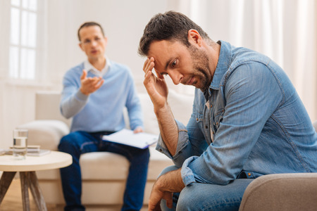 Life choice. Unhappy calm male patient looking down  touching his forehead  and sitting against the blurred backgroundの写真素材