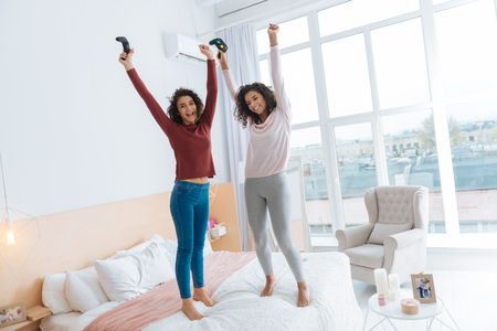 No time for worries. Full length shot of two curly haired ladies in casual beaming while holding video game consoles and jumping high on a bed.の写真素材