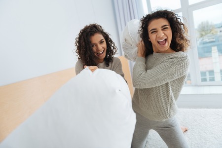 No time for worries. Extremely happy young ladies looking into the camera while sitting on a bed and getting ready for a pillow fight.の写真素材