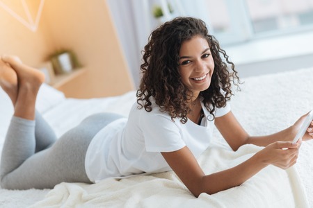 Such a lovely day. Radiant curly haired girl grinning broadly into the camera while relaxing on a bed and working on a touchpad.の写真素材