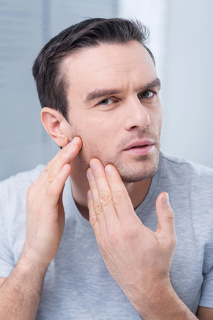 Something wrong. Calm serious young man touching his face and worrying about it while staring at the cameraの写真素材