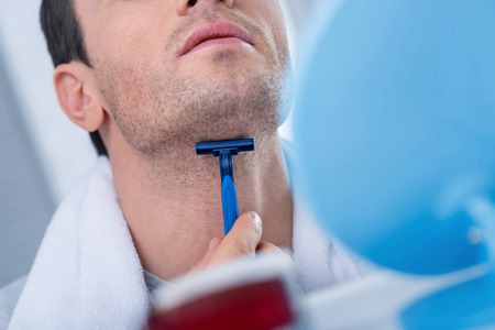 Accurate work. Close up of blue modern daily razor hold by man hands while he shaving his neck and standing in front of mirrorの写真素材