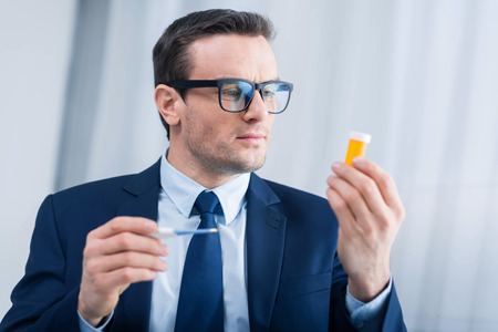 Interesting bottle. Thoughtful earnest confident man holding yellow bottle while examining it and wearing glassesの写真素材