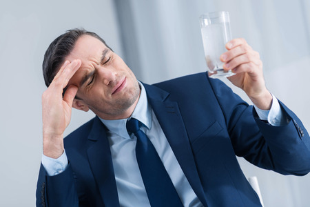 Headache driving crazy. Sad brunette young man closing his eyes while carrying glass of water and touching his foreheadの写真素材